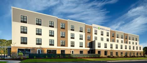 A multi-story hotel with a modern design, featuring a mix of brick and white exterior walls, large windows, and a sign indicating it is a home/suites hotel.