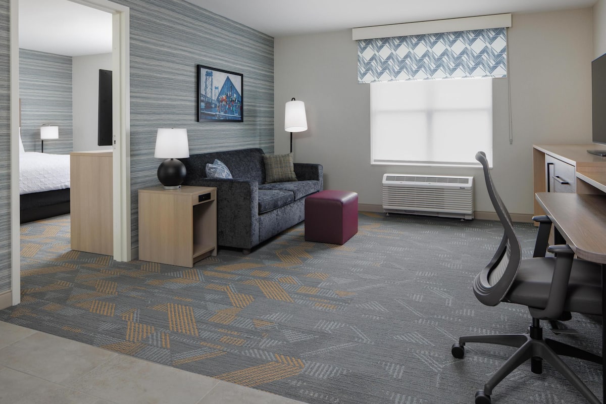 A hotel room with a bed, a sofa, a desk, a chair, a lamp, a heater, and a window with patterned curtains.
