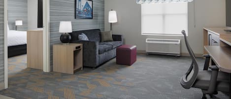 A hotel room with a bed, a sofa, a desk, a chair, a lamp, a heater, and a window with patterned curtains.