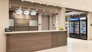 A hotel reception area with a modern design, featuring a reception desk, a sign for 'Home 2 Suites by Hilton', and a 'Hilton Honors' sign. There are two refrigerators in the background, one labeled 'COLDER'.