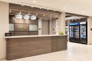 A hotel reception area with a modern design, featuring a reception desk, a sign for 'Home 2 Suites by Hilton', and a 'Hilton Honors' sign. There are two refrigerators in the background, one labeled 'COLDER'. - Home2 Suites By Hilton Kittery (Kittery)