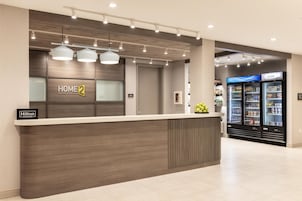 Zona de recepción de un hotel con diseño moderno, que incluye un mostrador de recepción, un letrero de 'Home 2 Suites by Hilton' y un letrero de 'Hilton Honors'. Se observan dos refrigeradores al fondo, uno etiquetado como 'COLDER'.