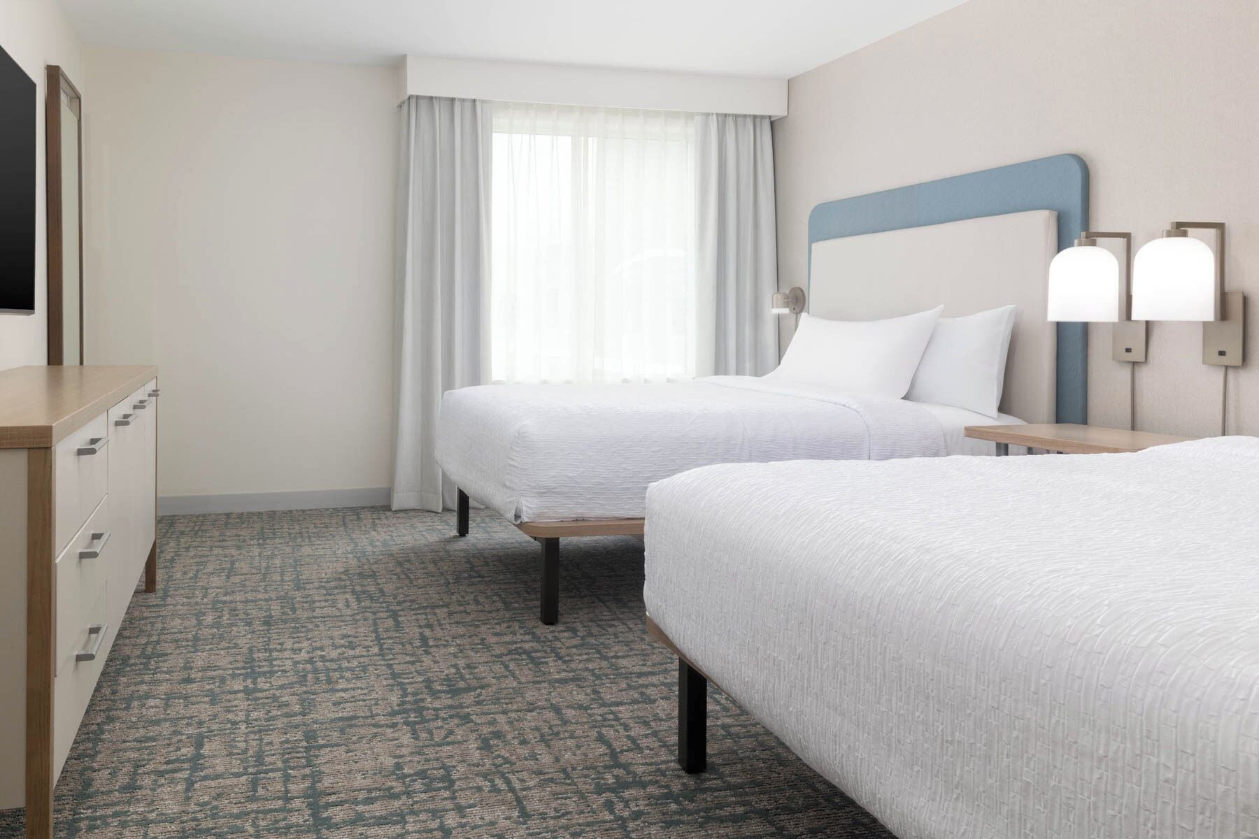A hotel room with two beds, a desk, a chair, a window with curtains, and wall-mounted lights.