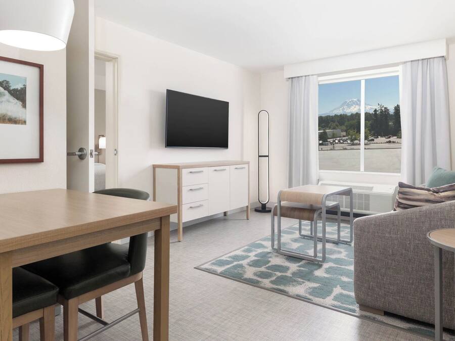 A modern hotel room with a dining area, a sofa, a TV, and a view of a mountain and water.