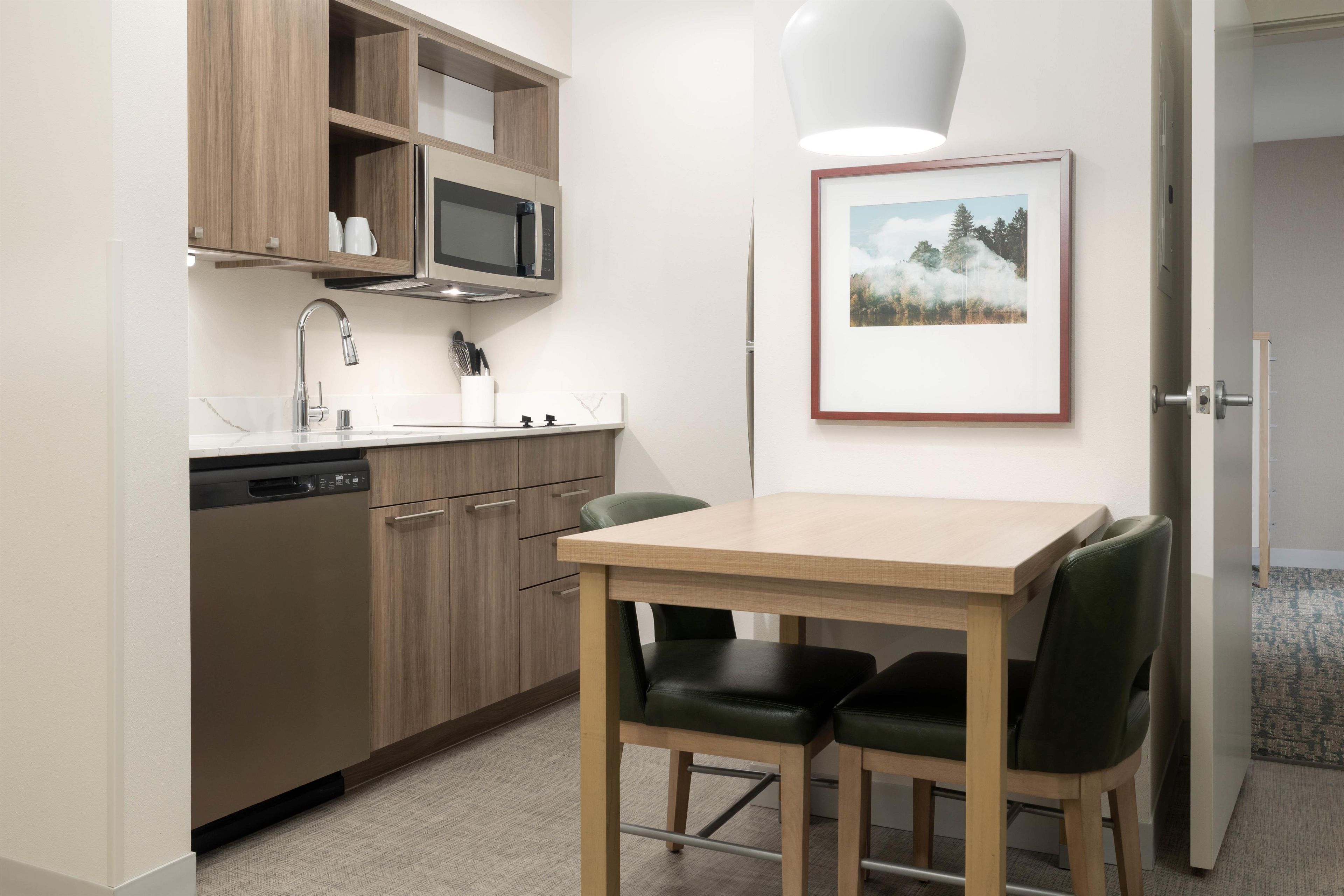A compact kitchen with a microwave, sink, and dishwasher. There is a framed picture on the wall and a table with chairs.