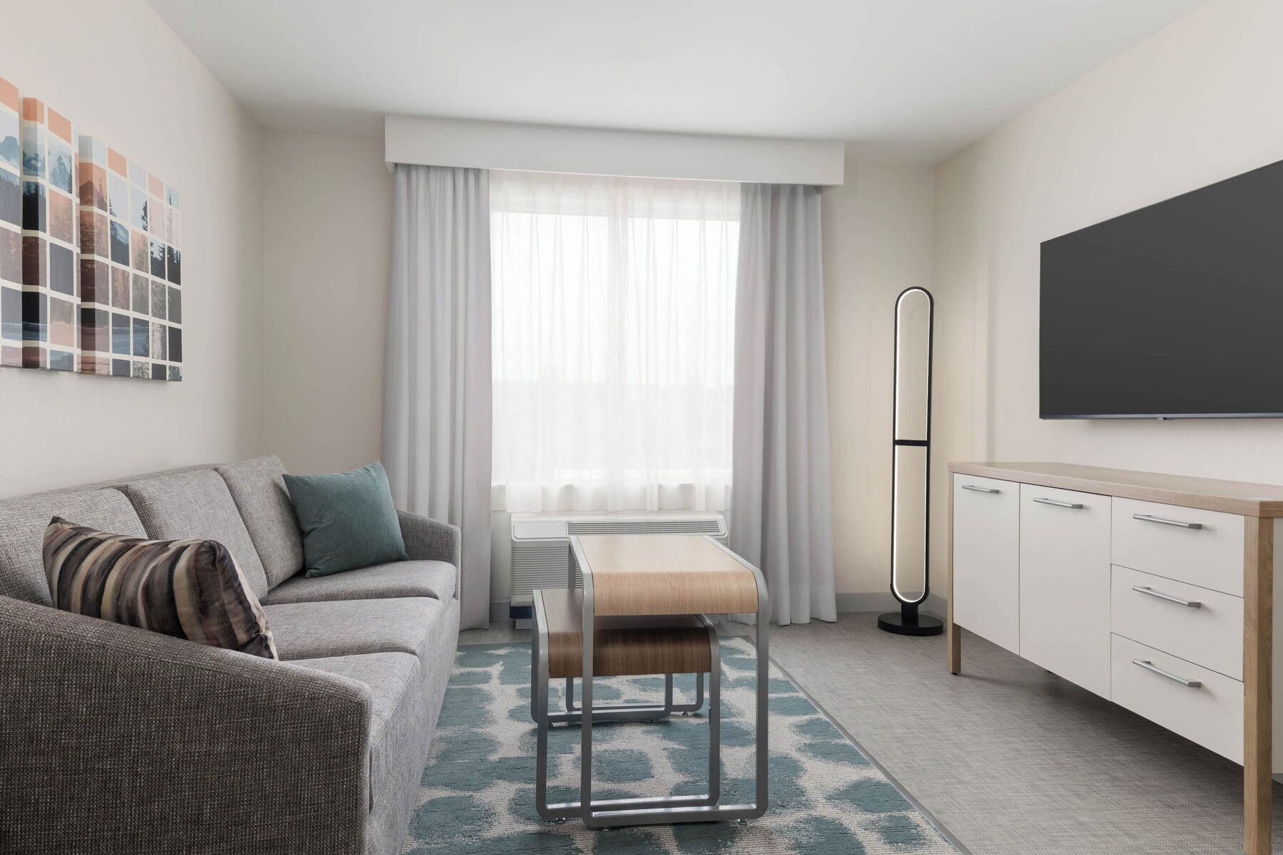 A modern hotel room with a grey sofa, a wooden bench, a white cabinet, and a large TV on the wall.