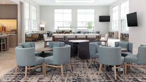 A modern dining area with round tables and grey upholstered chairs, large windows, and a flat-screen TV mounted on the wall.