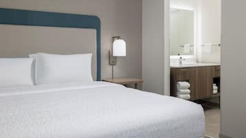 Una habitación de hotel con cama, mesita de noche, lámpara de pared, baño con lavamanos y toallero.