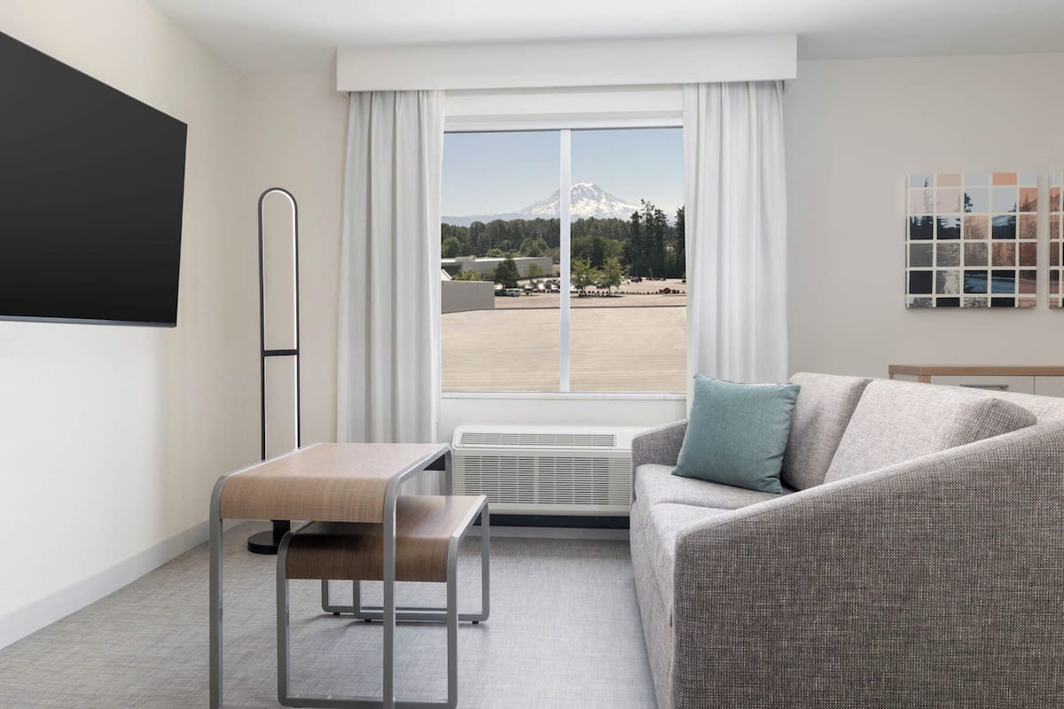 A modern hotel room with a large window, a sofa, a small table, and a standing lamp.