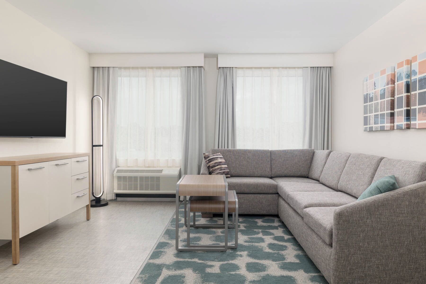 Ein modernes Wohnzimmer mit grauem Sofa, einem kleinen Holztisch, einem Fernseher an der Wand und einem großen Fenster mit transparenten Vorhängen.