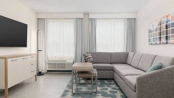 Een moderne woonkamer met een grijze bank, een klein houten tafeltje, een televisie aan de muur en een groot raam met dunne gordijnen.