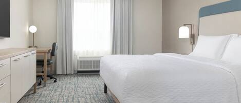 Ett hotellrum med en säng, ett skrivbord, en stol och ett stort fönster med gardiner.