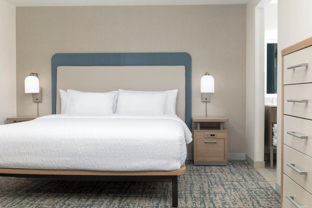 A neatly made bed with a wooden frame and white bedding, flanked by two bedside tables with lamps, in a room with a patterned carpet and a neutral-toned wall.
