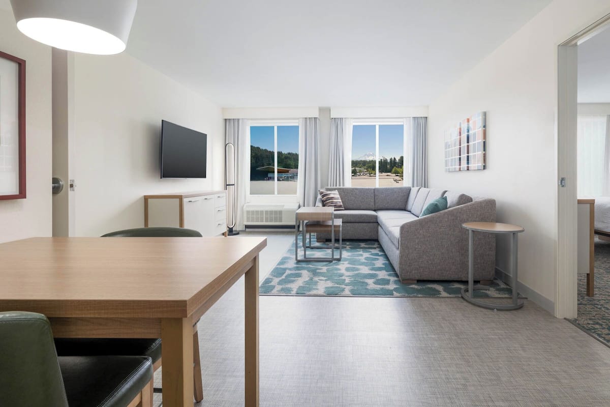A modern hotel room with a wooden dining table, a grey sofa, a small round table, and a TV mounted on the wall.