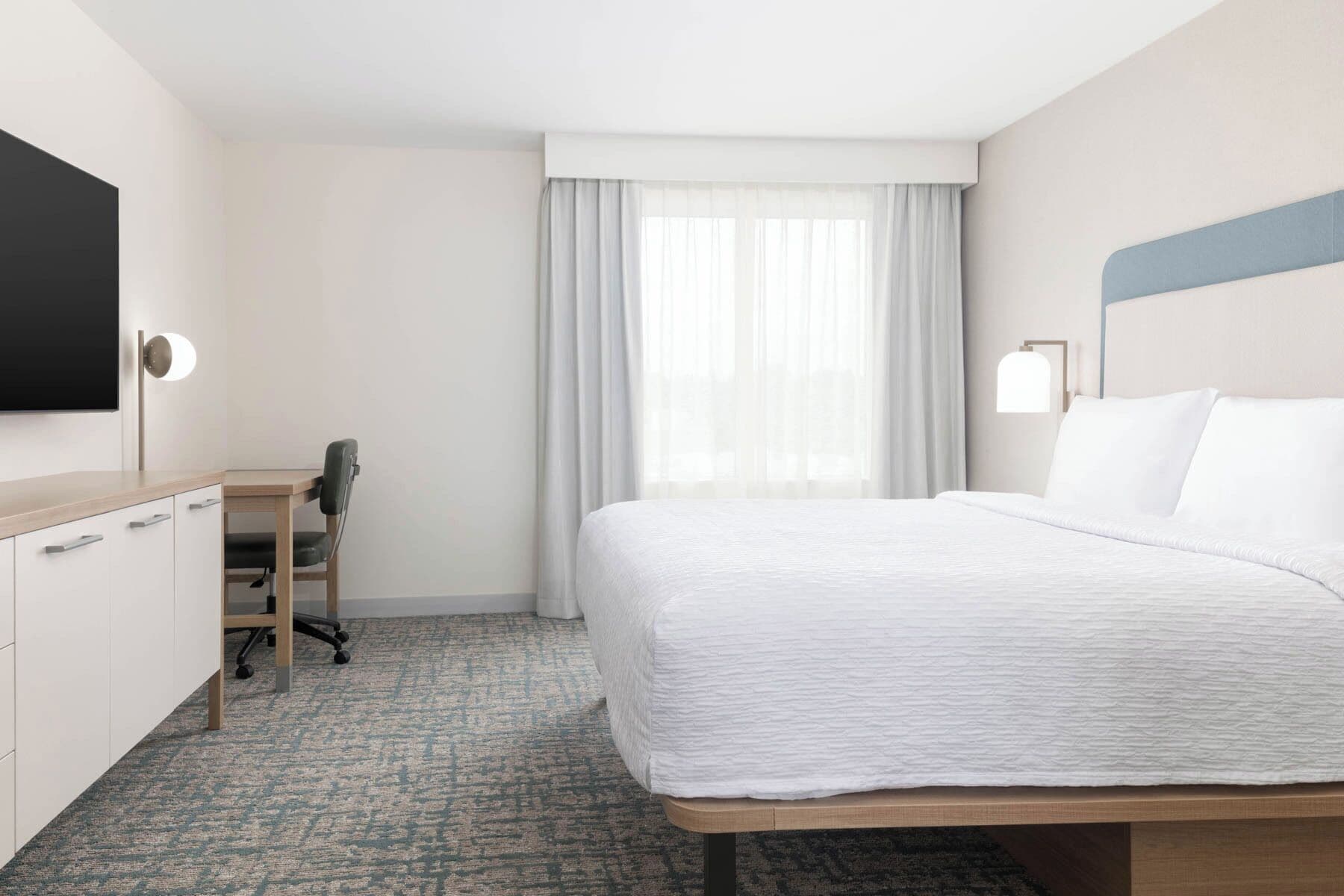 A hotel room with a bed, a desk with a chair, a TV, and a window with curtains.