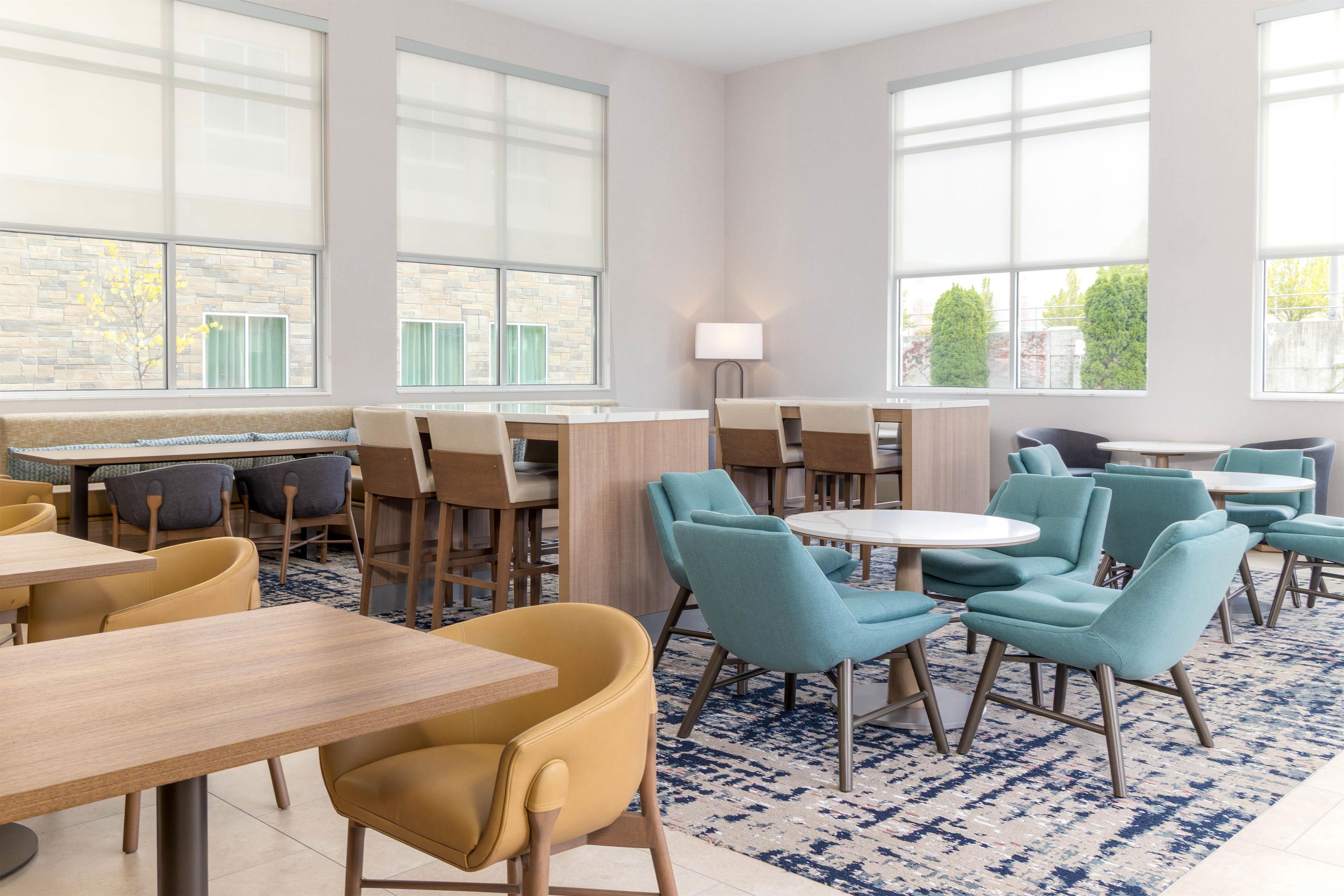 A hotel dining area with multiple tables and chairs, large windows, and a patterned carpet.