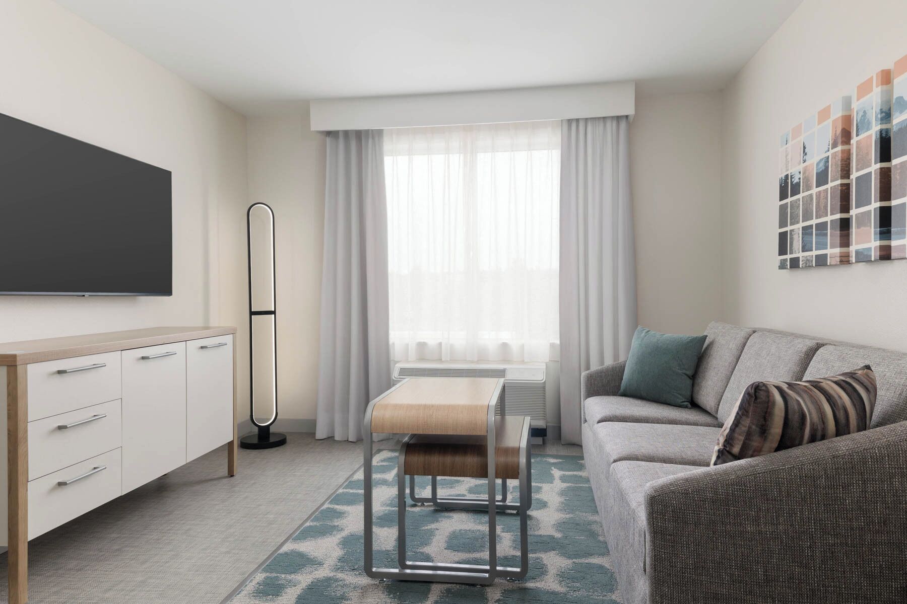 Una habitación de hotel moderna con una televisión de pantalla plana, un sofá, una mesita pequeña y un ventanal con cortinas translúcidas.