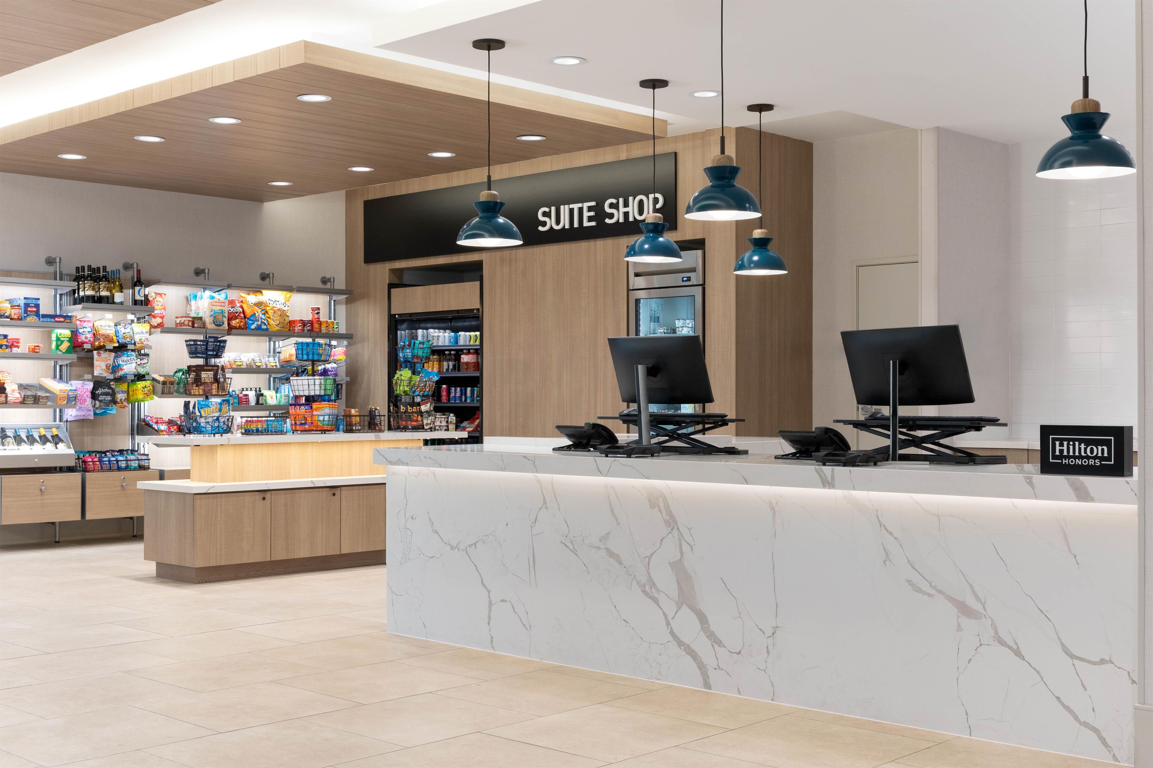 A hotel suite shop with a marble counter, overhead lighting, and various food and beverage products on shelves.