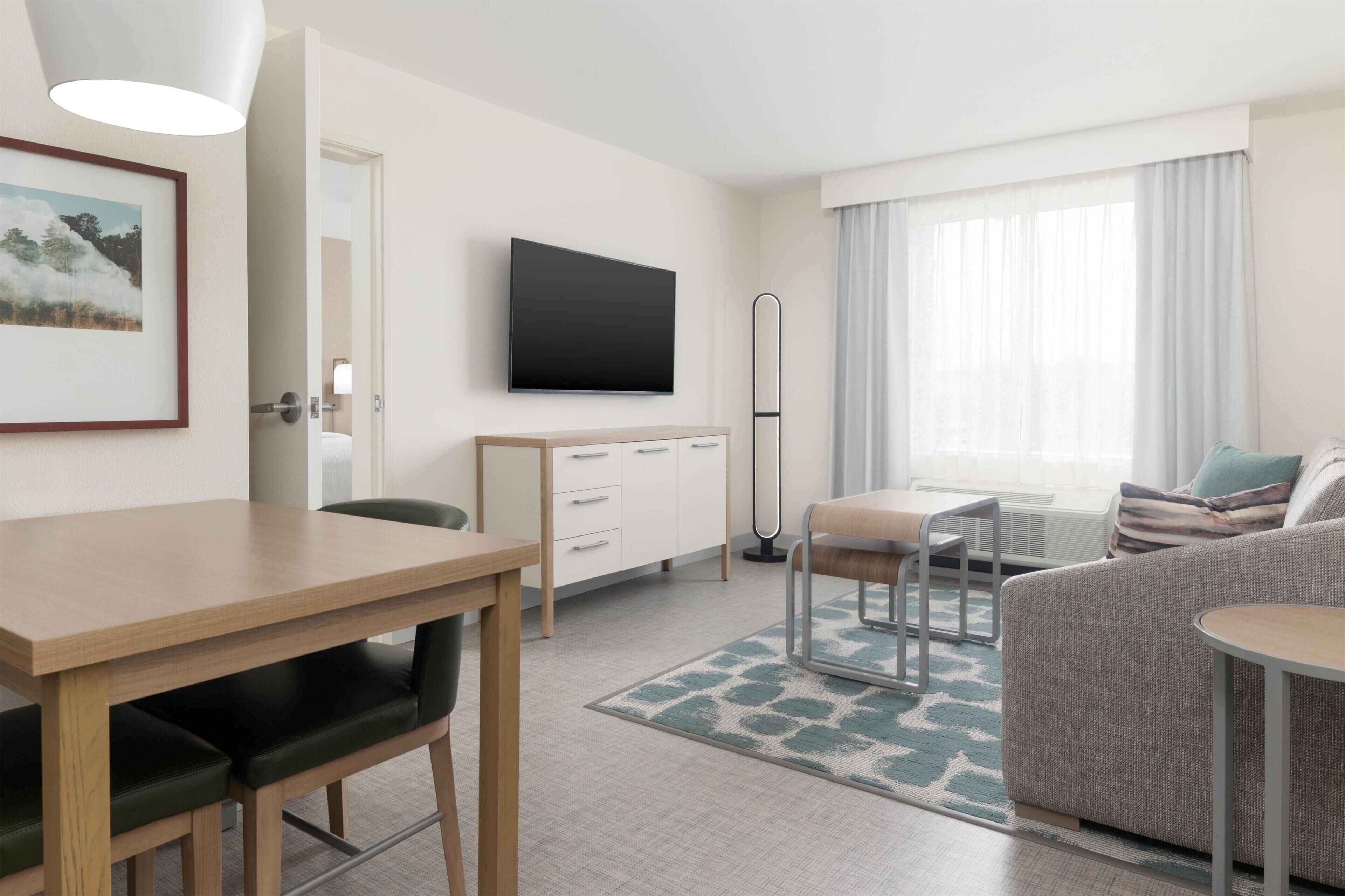 A modern hotel room with a flat-screen TV, a dining area with a table and chairs, a sofa, and a large window with curtains.