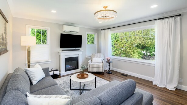 A modern living room with a grey sofa, a fireplace, a television, and large windows with curtains.