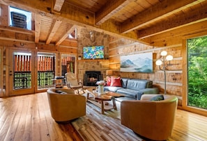 A log cabin interior with a stone fireplace, wooden furniture, and a large window overlooking greenery.