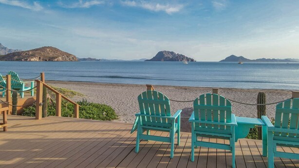 Una terraza de madera con sillas turquesas que da a una playa con montañas lejanas y un cielo despejado.