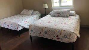 A room with two single beds, each with patterned bedding, a bedside table with a lamp, and a window with a view of the outside.