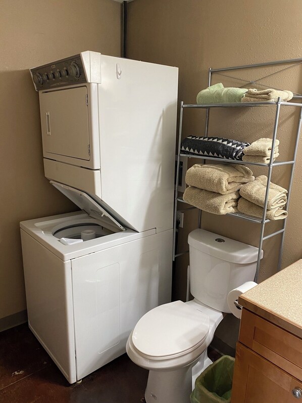 Un espace buanderie compact comprenant un lave-linge et un sèche-linge, des toilettes et une étagère en métal où sont rangées les serviettes.