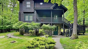 Une cabane en rondins à deux étages, dotée d’un balcon, entourée d’une végétation luxuriante et bordée d’un sentier en pierre.
