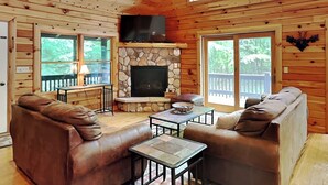 L’intérieur chaleureux d’un chalet avec une cheminée en pierre, des canapés en cuir et de grandes fenêtres donnant sur le paysage extérieur.