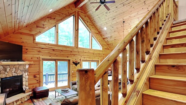 Un intérieur de cabane spacieux, doté d’une cheminée en pierre, d’un escalier en bois et de grandes fenêtres offrant une vue sur l’extérieur.
