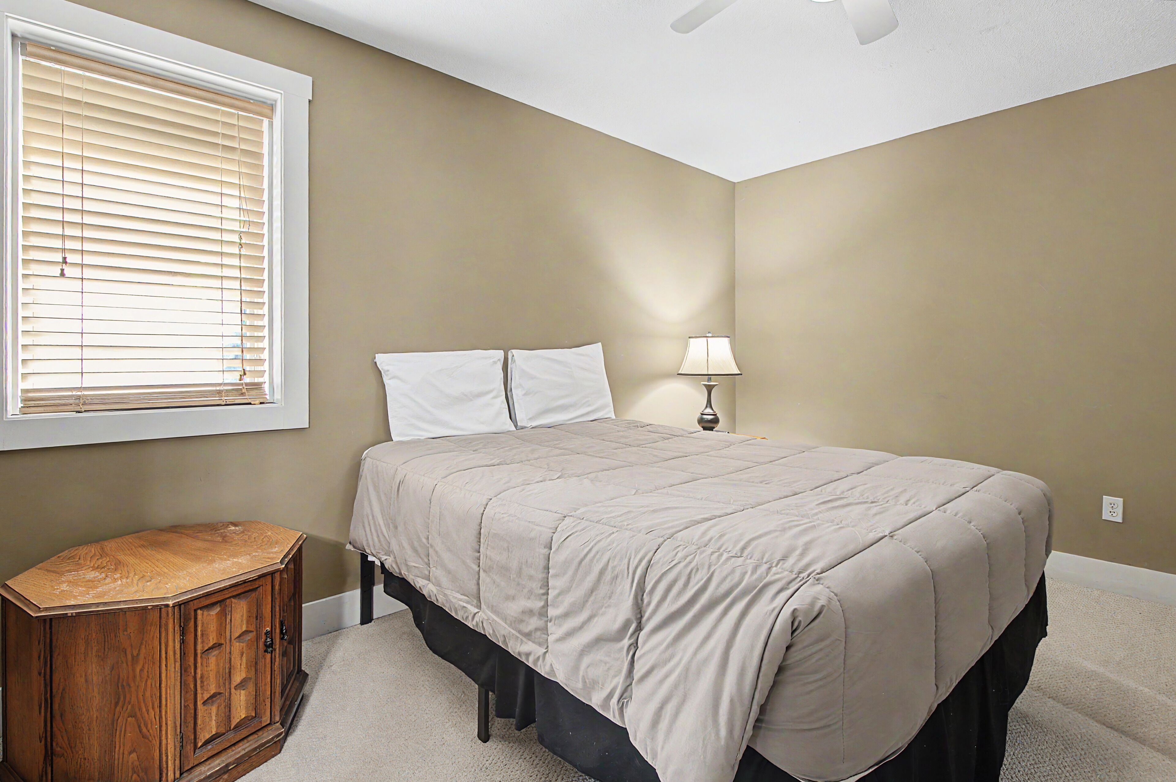 A bedroom with a bed, a nightstand, a lamp, a window with blinds, and a ceiling fan.