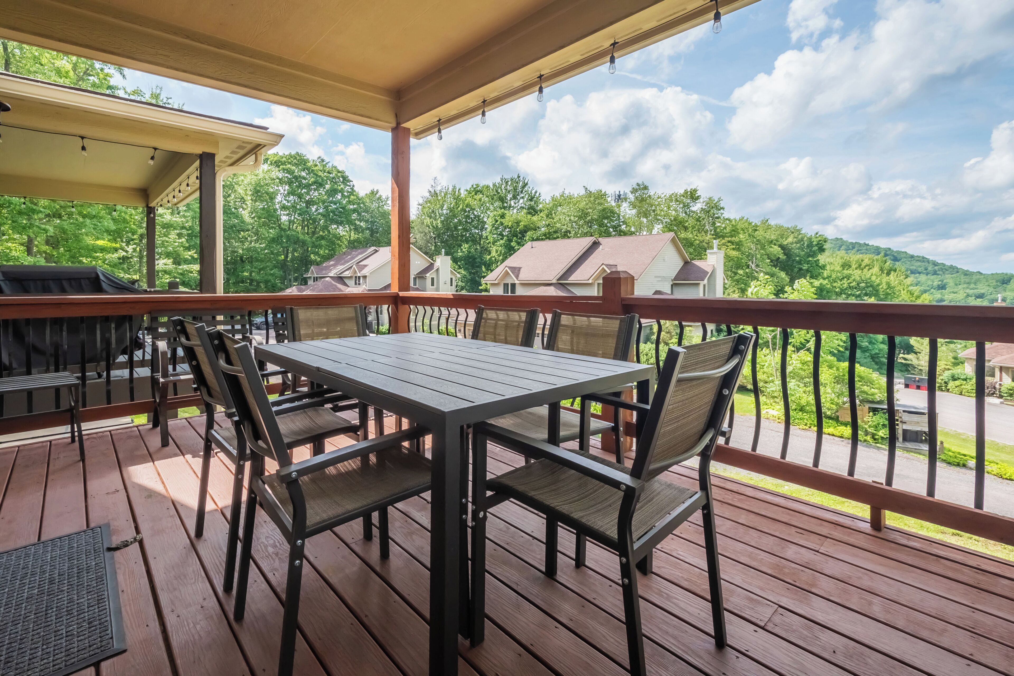 A covered outdoor deck with a table and chairs, overlooking a residential area and hills.