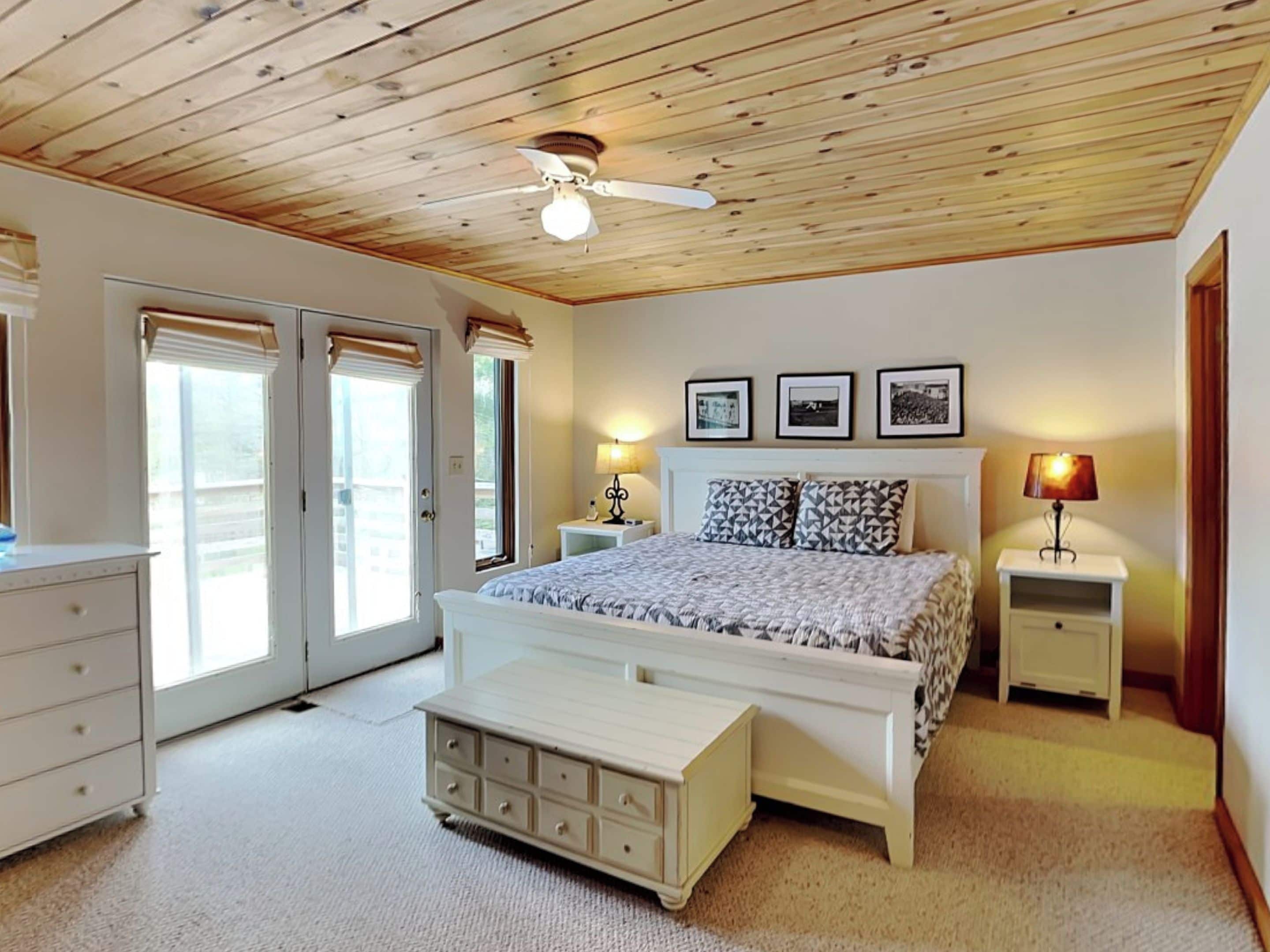 A bedroom with a wooden ceiling, a bed with patterned bedding, bedside tables with lamps, and a dresser.