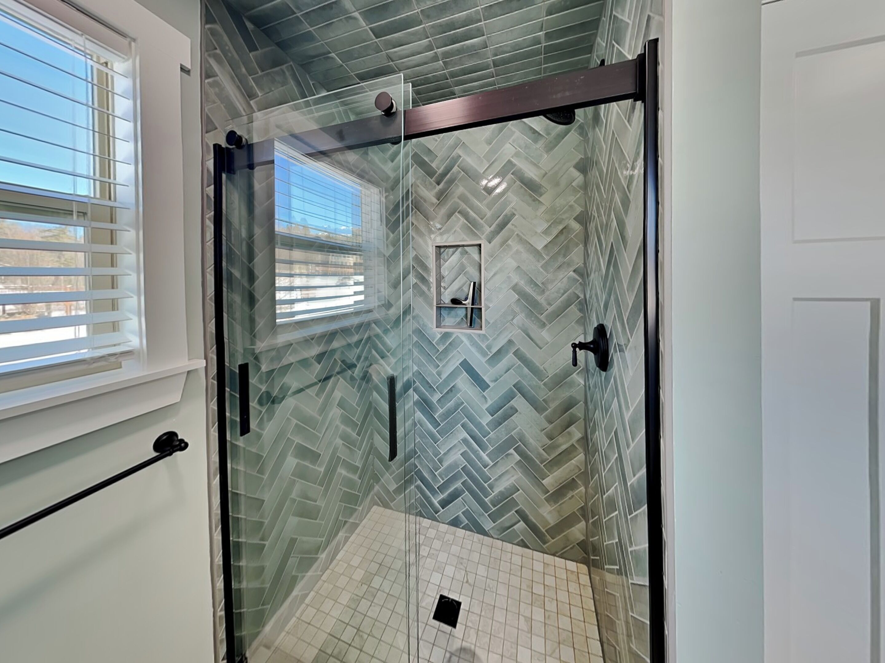 A glass-enclosed shower with a herringbone tile pattern, a window with blinds, and a door with a handle.