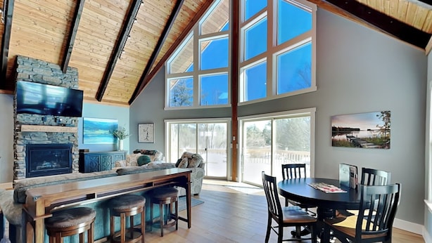 A spacious living area with a stone fireplace, a large window, and a dining table with chairs. - As You Lake It by Taylor Made Deep Creek (Oakland)