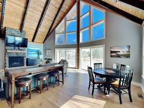 A spacious living area with a stone fireplace, a large window, and a dining table with chairs. - As You Lake It by Taylor Made Deep Creek (Oakland)