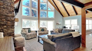 A spacious living room with a high ceiling, stone wall, and large windows offering a view of trees. - As You Lake It by Taylor Made Deep Creek (Oakland)