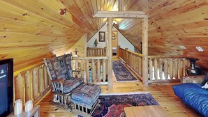 A wooden interior with a loft bedroom, a balcony, and a seating area with patterned cushions.