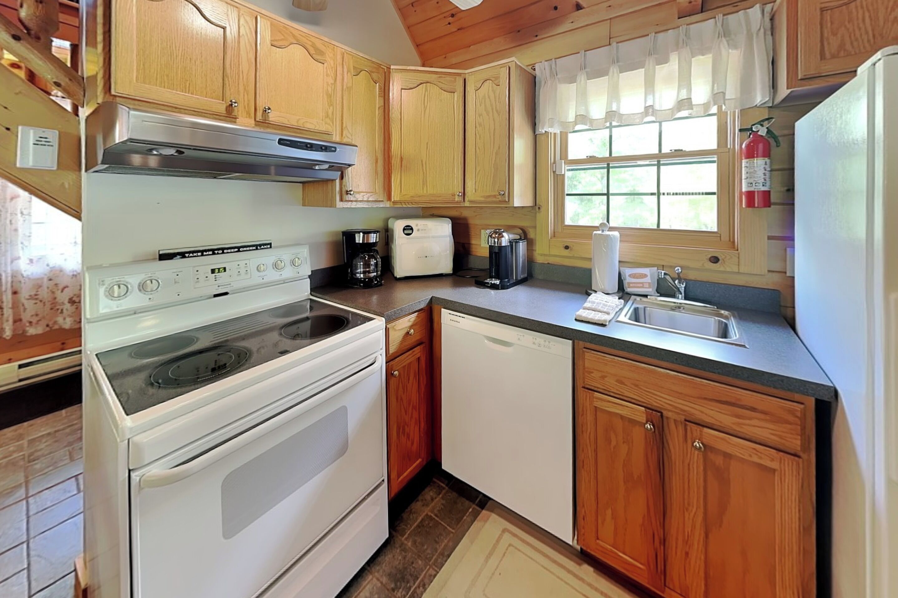 A kitchen with wooden cabinets, a stove, a dishwasher, a coffee maker, and a window with curtains.