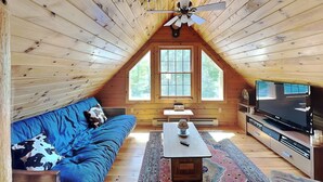 An attic bedroom with a wooden ceiling, a ceiling fan, a large window, a sofa, a television, and a coffee table.