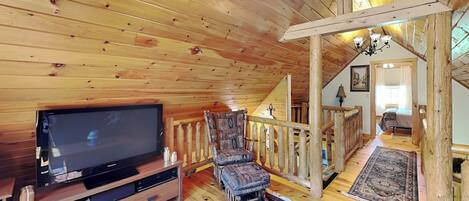 A wooden interior with a television, a patterned chair, a wooden staircase, and a chandelier.