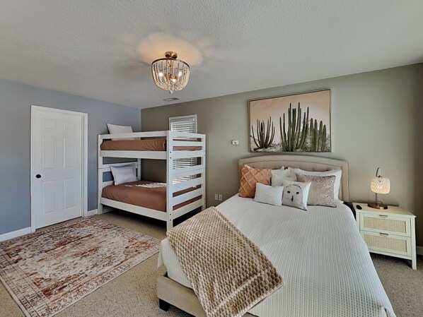 A bedroom with bunk beds, a large bed, a nightstand, a lamp, a door, and a cactus painting on the wall. - Dacha Verde by Taylor Made Deep Creek (Swanton)