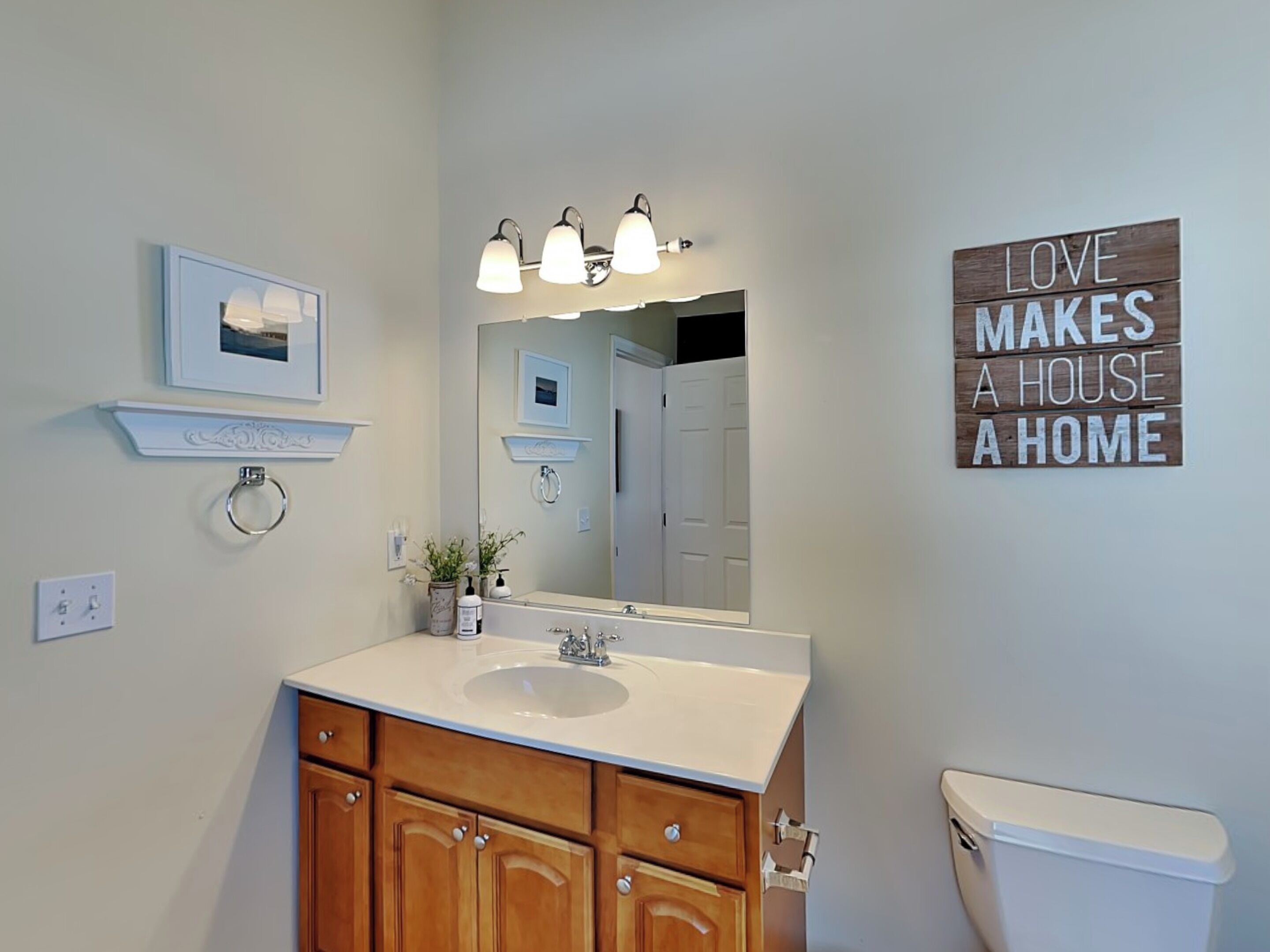 A bathroom with a white sink, wooden cabinets, a mirror, and a sign saying "LOVE MAKES A HOUSE A HOME".