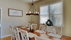 A dining room with a wooden table, white chairs, a chandelier, and a window with blinds. - Dacha Verde by Taylor Made Deep Creek (Swanton)