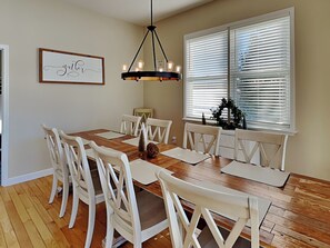A dining room with a wooden table, white chairs, a chandelier, and a window with blinds. - Dacha Verde by Taylor Made Deep Creek (Swanton)