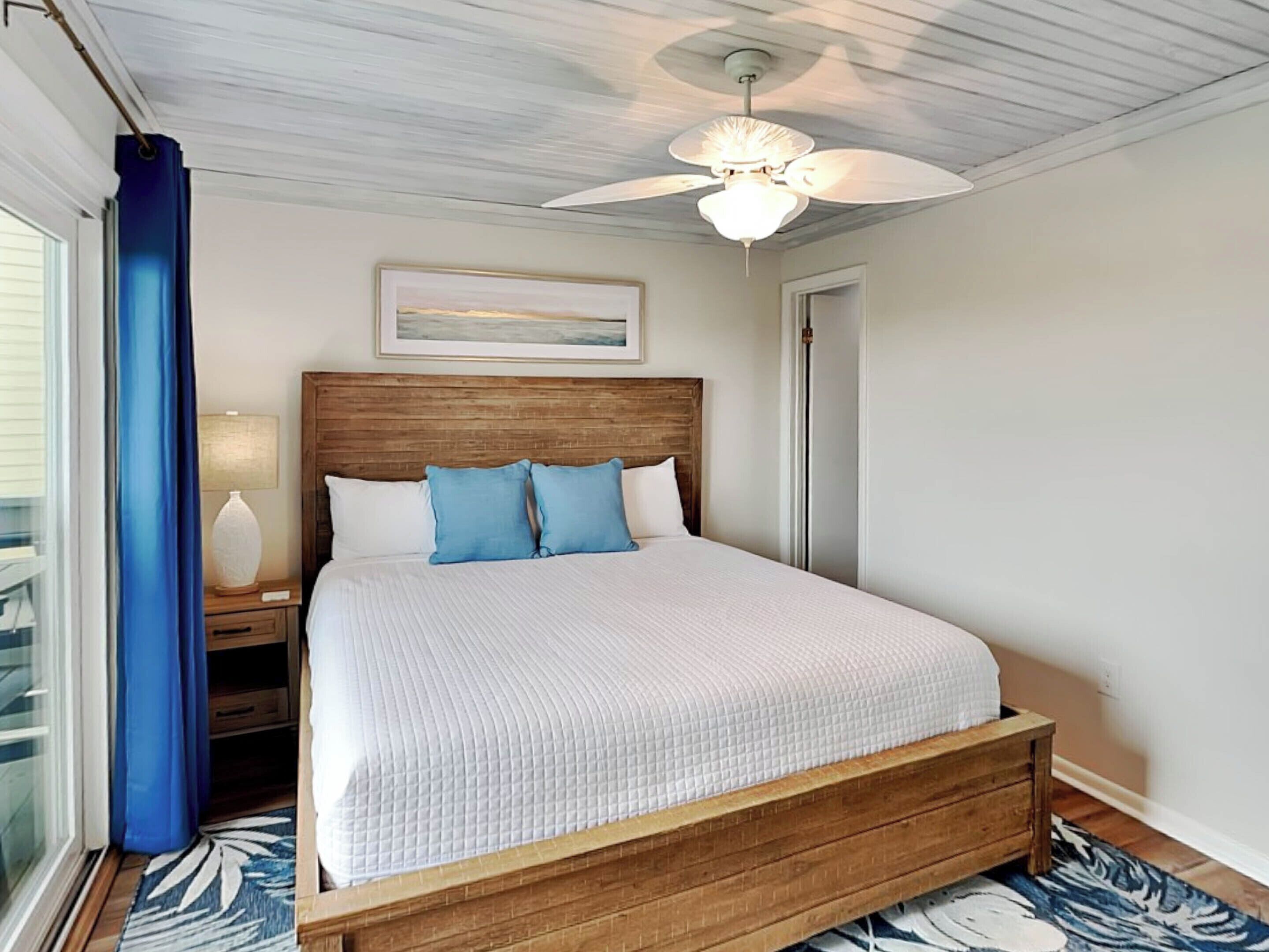 A bedroom with a wooden bed frame, white bedding, a ceiling fan, a nightstand with a lamp, and a window with blue curtains.