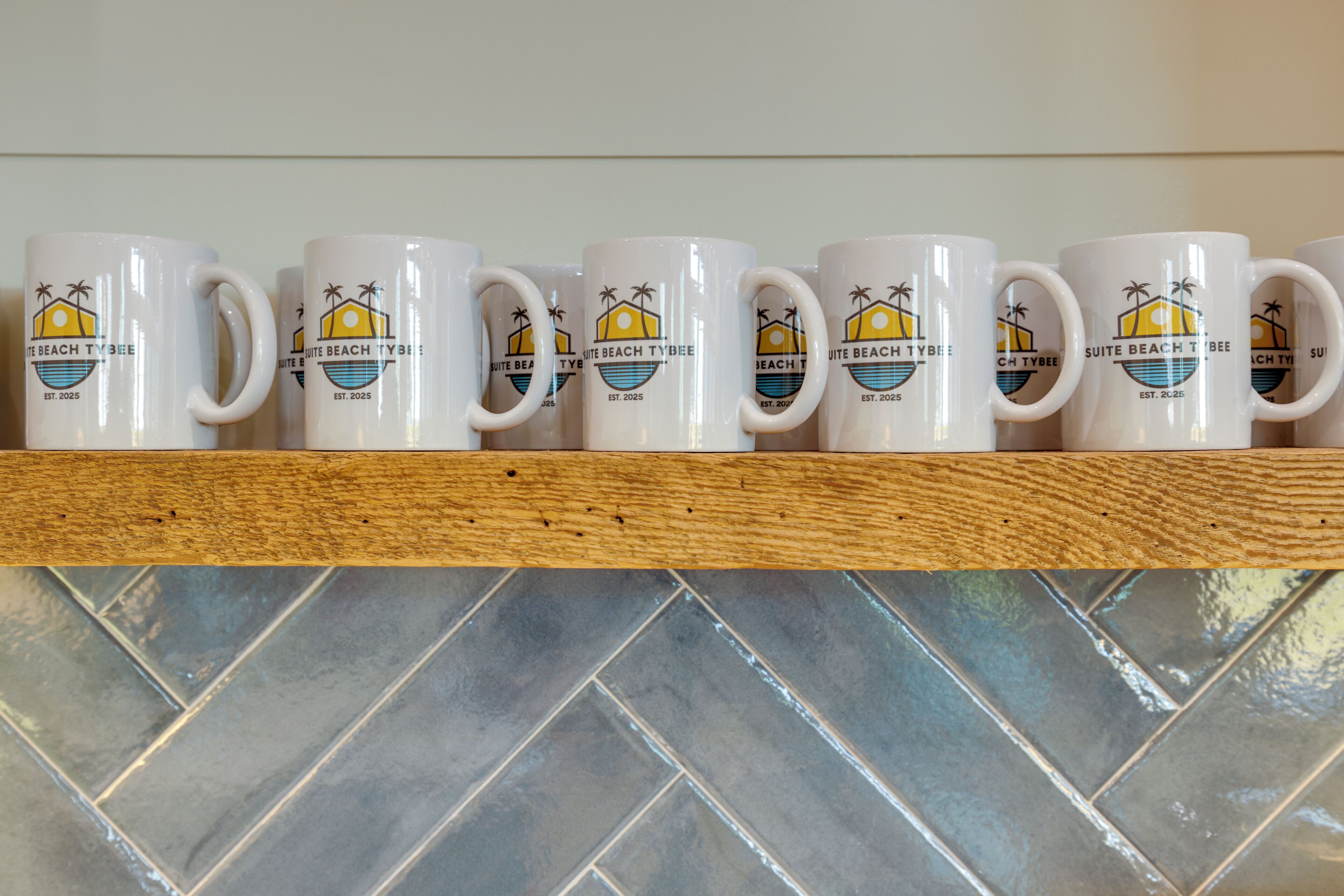 A row of white mugs with a beach design and the text "SUITE BEACH TYBEE EST. 2025" on a wooden shelf.