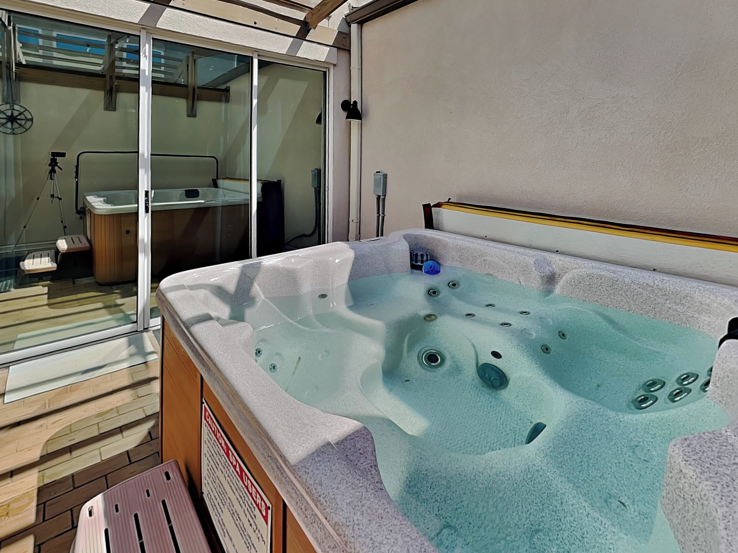 Un bain à remous doté d’une porte en verre donnant sur une salle de bain, une terrasse en bois et un panneau d’avertissement à l’attention des utilisateurs du spa.