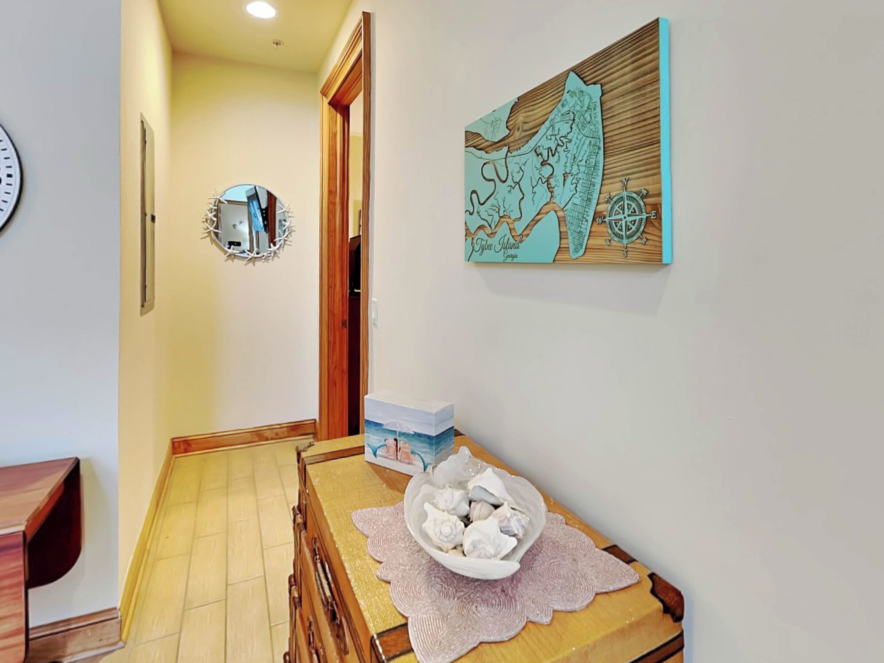 A hallway with a wooden floor, a dining table with a white bowl and a box, a wall clock, and a map artwork.